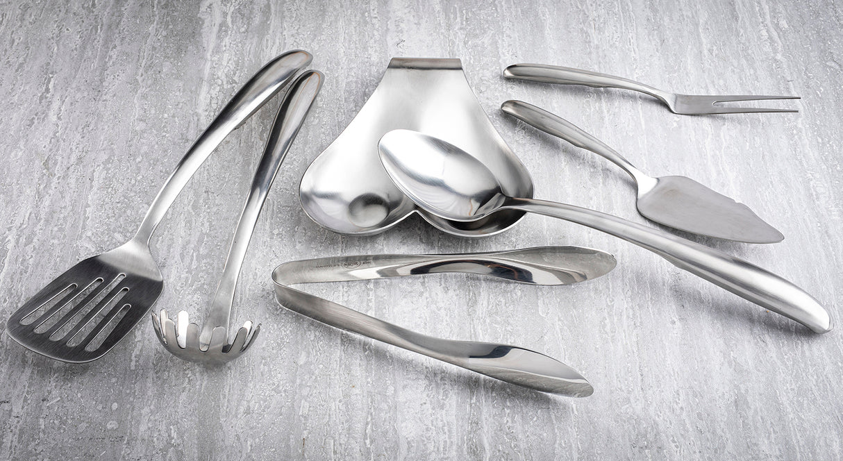 A variety of stainless steel kitchen utensils from the Dalton Series Utensil Set, including tongs, serving spoons, and a slotted spatula, are arranged on a light gray textured surface. The utensils are clean and reflective.