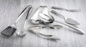 A variety of stainless steel kitchen utensils from the Dalton Series Utensil Set, including tongs, serving spoons, and a slotted spatula, are arranged on a light gray textured surface. The utensils are clean and reflective.