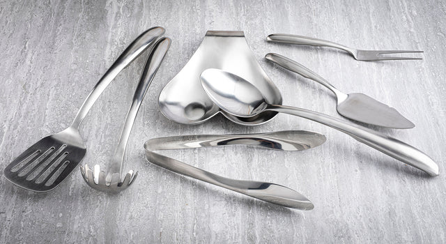 A variety of stainless steel kitchen utensils from the Dalton Series Utensil Set, including tongs, serving spoons, and a slotted spatula, are arranged on a light gray textured surface. The utensils are clean and reflective.