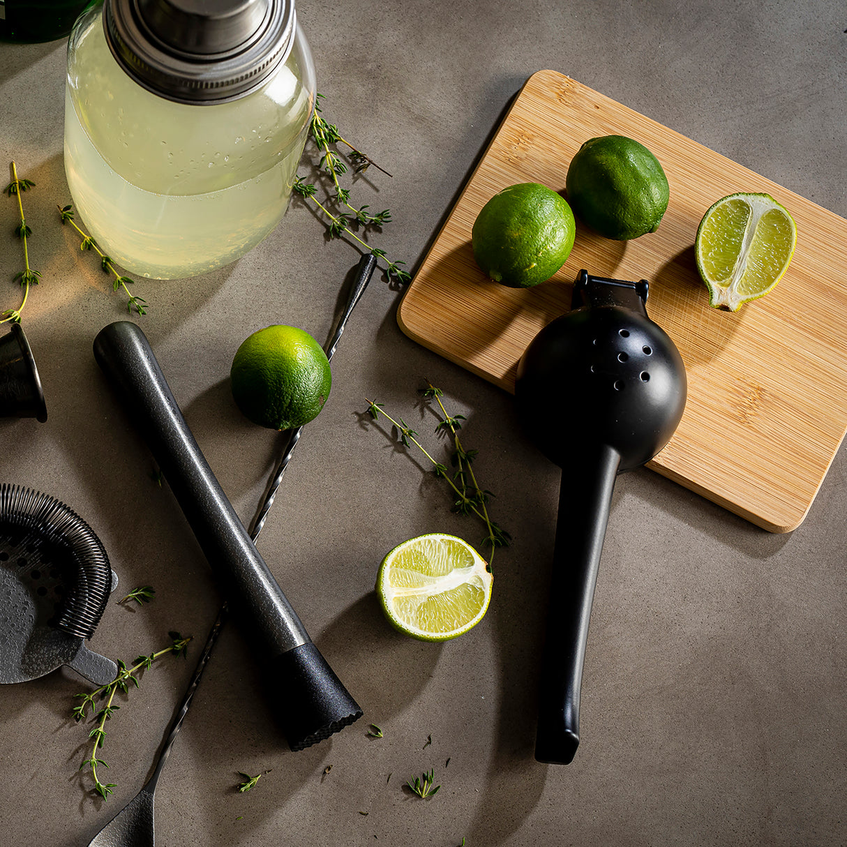 A cocktail-making scene on a gray countertop: a shaker, strainer, muddler, and bar spoon, with fresh limes—some whole, some halved—on a wooden cutting board. Sprigs of thyme and a TableCraft coated aluminum alloy juicer complete the setup.