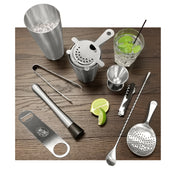 A Premium Barware Set on a wooden surface: stainless steel cocktail shaker with ice, strainer, tongs, jigger, muddler, bottle opener, mixing spoons, corkscrew, sliced lime, and a glass with a green drink, ice, lime, and black straw.