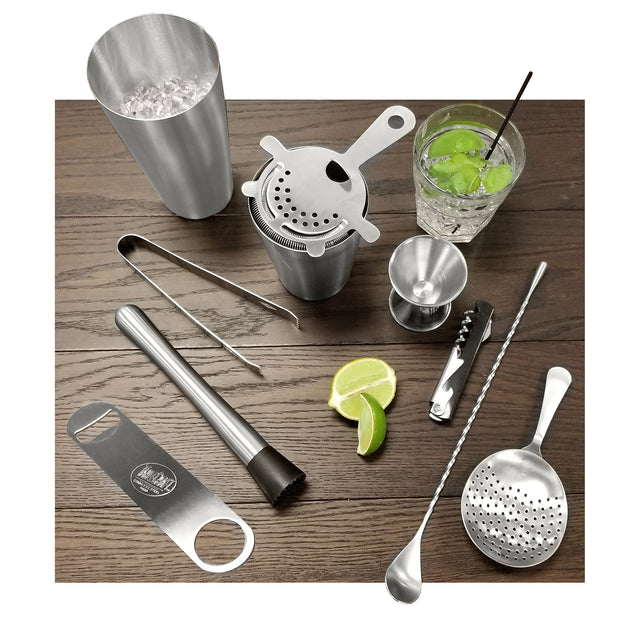A Premium Barware Set on a wooden surface: stainless steel cocktail shaker with ice, strainer, tongs, jigger, muddler, bottle opener, mixing spoons, corkscrew, sliced lime, and a glass with a green drink, ice, lime, and black straw.