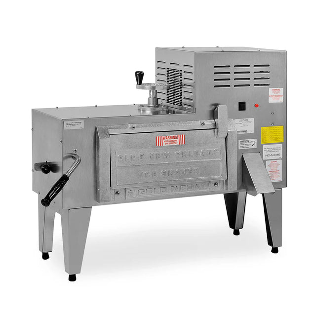 A commercial beignet fryer made of stainless steel with a boxy shape, labeled Olde New Orleans, stands on four legs beside a high volume ice shaver. It features a front-loading door, heat vents, control knobs, and instructional stickers on the side.