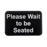 A rectangular black TableCraft 10901 self-adhesive sign with rounded corners displays the white text: “Please Wait to be Seated.” The message is centered, easy to read, and set against a plain background.