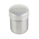 A dishwasher safe stainless steel shaker with a translucent plastic lid labeled Cocoa. The lid has small holes for sprinkling and fits snugly on the round, metallic container, which reflects light from its smooth surface. The background is plain white.
