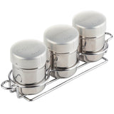 Three stainless steel spice shakers with white lids labeled Cocoa, Cinnamon, and Nutmeg sit in a chrome-plated rack. The cylindrical shaker set is arranged in a row, with each label printed on the top of the lid.