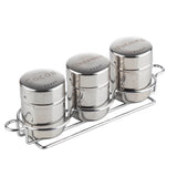 Three shiny stainless steel shakers labeled “Cocoa,” “Cinnamon,” and “Nutmeg” sit side by side in a chrome-plated rack. The shaker set features lids with small holes for sprinkling, and each container is clearly engraved.