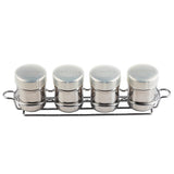 Four small stainless steel spice shakers with white lids, labeled Cinnamon, Vanilla, Cocoa, and Nutmeg, are neatly arranged in a chrome-plated rack with handles on both ends, set against a plain white background.