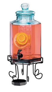 A 2 gallon beverage dispenser with a lid holds pink liquid, ice, and lemon slices. It sits on a decorative black metal stand with curly legs and features a black spigot above a small drip tray.