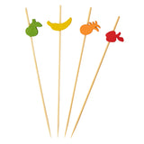 Four wooden decorative picks, each topped with a colored fruit-shaped cutout—green pear, yellow banana, orange pineapple, and red apple—are arranged in a row on a white background. Ideal for parties, these are TableCraft 11204 bamboo pinewood picks.