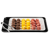 A black rectangular platter holds a white plate with three neat rows of colorful truffles: brown (left), yellow (center), and pink (right). Each row has five truffles, with silver tongs on the Sierra Collection serving platter’s edge. The background is white.
