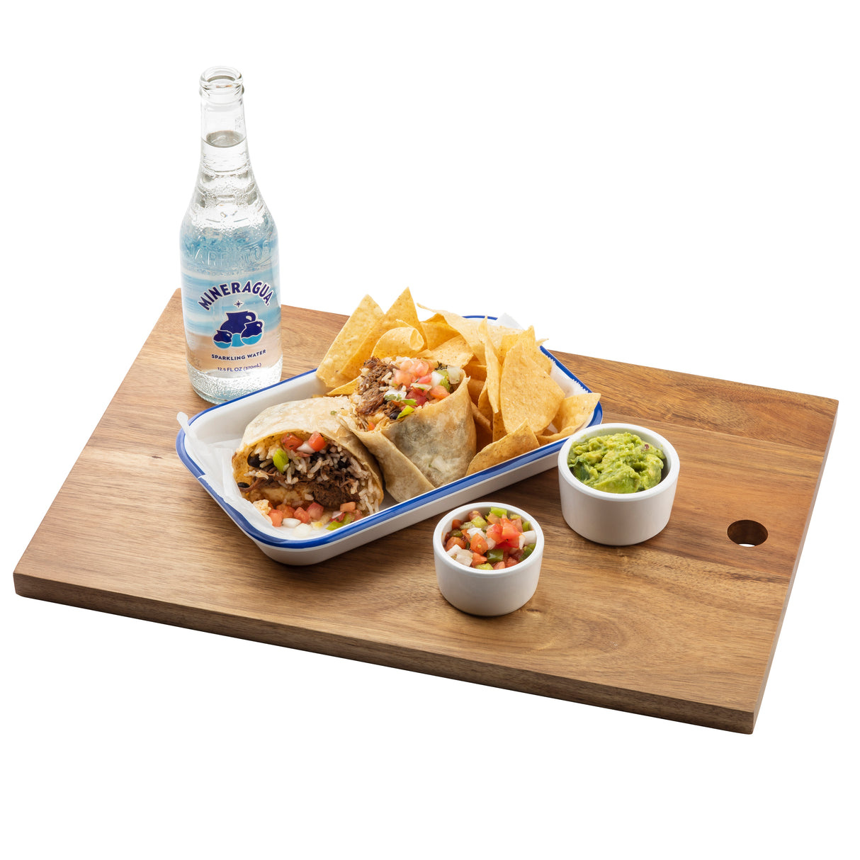 A TableCraft serving board holds a metal tray with a burrito cut in half, tortilla chips, guacamole and salsa in ramekins, plus a bottle of sparkling water. The burrito is filled with rice, beans, lettuce, tomatoes, and possibly meat.