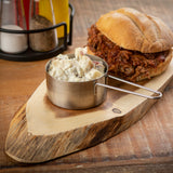 A metal measuring cup filled with creamy potato salad sits on a TableCraft Products Acacia Serving Board. Behind it rests a pulled pork sandwich on a crusty bun, with salt, pepper, and condiments in the background on a wooden table.
