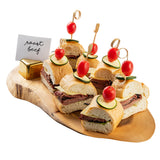 A TableCraft Products Acacia Serving Board displays roast beef sandwiches, each topped with cucumber and cherry tomato skewers. A small "roast beef" sign sits at the back of the wood serving board against a white background.