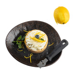 A lemon tart topped with whipped cream, a blueberry, and lemon zest sits on a dark Lunara Collection serving plate with a mint sprig. A zester tool and whole lemon rest nearby; scattered zest decorates the plate against a white background.