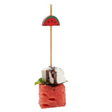 A skewer made from natural bamboo holds a watermelon slice, basil leaf, and feta cube drizzled with balsamic glaze. Topped with a decorative pick shaped like a watermelon slice, this TableCraft accessory pops against a white background.