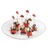 Six appetizer skewers with watermelon cubes, basil leaves, and feta cheese topped with balsamic glaze are arranged on a white plate. Each skewer uses Natural Bamboo Decorative Picks by TableCraft with a small watermelon image at the top.