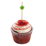 A red velvet cupcake with white cream cheese frosting, topped with red crumbs. A TableCraft Decorative Pick made from natural bamboo, featuring a small green paper star, is inserted in the center. The cupcake sits in a red and white liner on a white background.