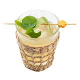 A glass of light yellow drink sits in a woven wicker holder. Garnished with a green leaf and curly peel on decorative picks from TableCraft Products, the drink features a yellow flower bead, set against a crisp white background.
