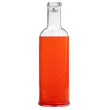 A clear glass TableCraft carafe, with a 34 oz. capacity, is filled with a bright orange or red carbonated liquid. Seen from the front, bubbles rise through the fizzy drink, and a transparent cap seals the narrow neck.