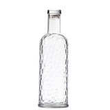 A clear glass TableCraft carafe with a tall neck and hammered, textured surface. This 34 oz bottle features a round base, smooth narrow rim, and faceted, dimpled design, standing upright against a plain white background.