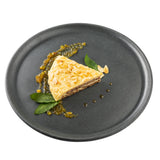 A slice of layered cake topped with almond flakes sits on a Terra Collection serving plate, garnished with mint leaves and a yellow fruit sauce swirl with small droplets, all set against a white background.