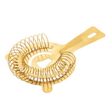 A gold-colored stainless steel cocktail strainer with a flat handle and large coiled spring encircling its base. The handle has a rectangular hole near the end, and prongs for resting the strainer on a shaker or glass.