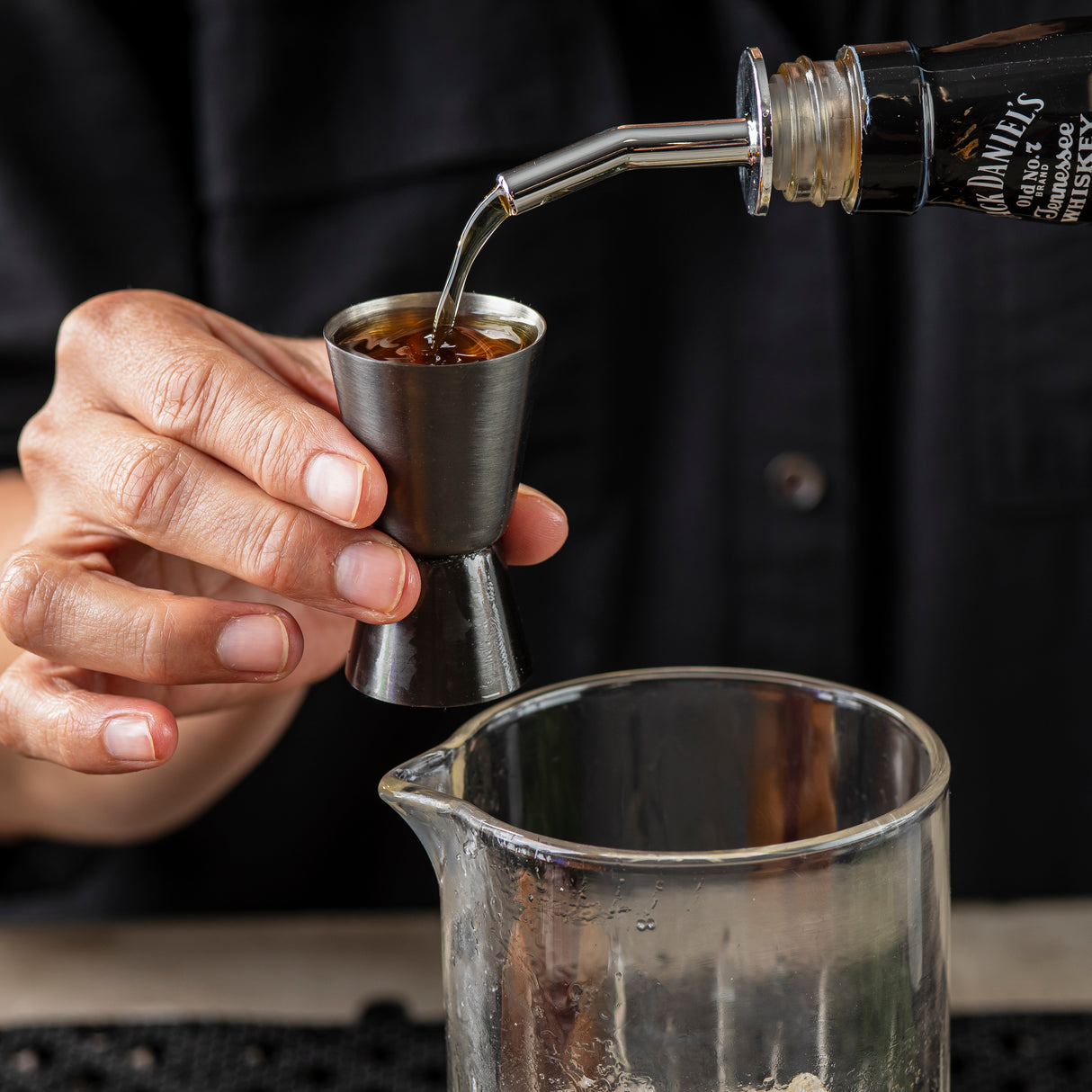 A person holds a stainless steel jigger while pouring whiskey from a Jack Daniel’s bottle into it above a clear glass mixing beaker. The person wears a dark shirt, and only their hands and torso are visible.