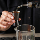 A person holds a stainless steel jigger while pouring whiskey from a Jack Daniel’s bottle into it above a clear glass mixing beaker. The person wears a dark shirt, and only their hands and torso are visible.