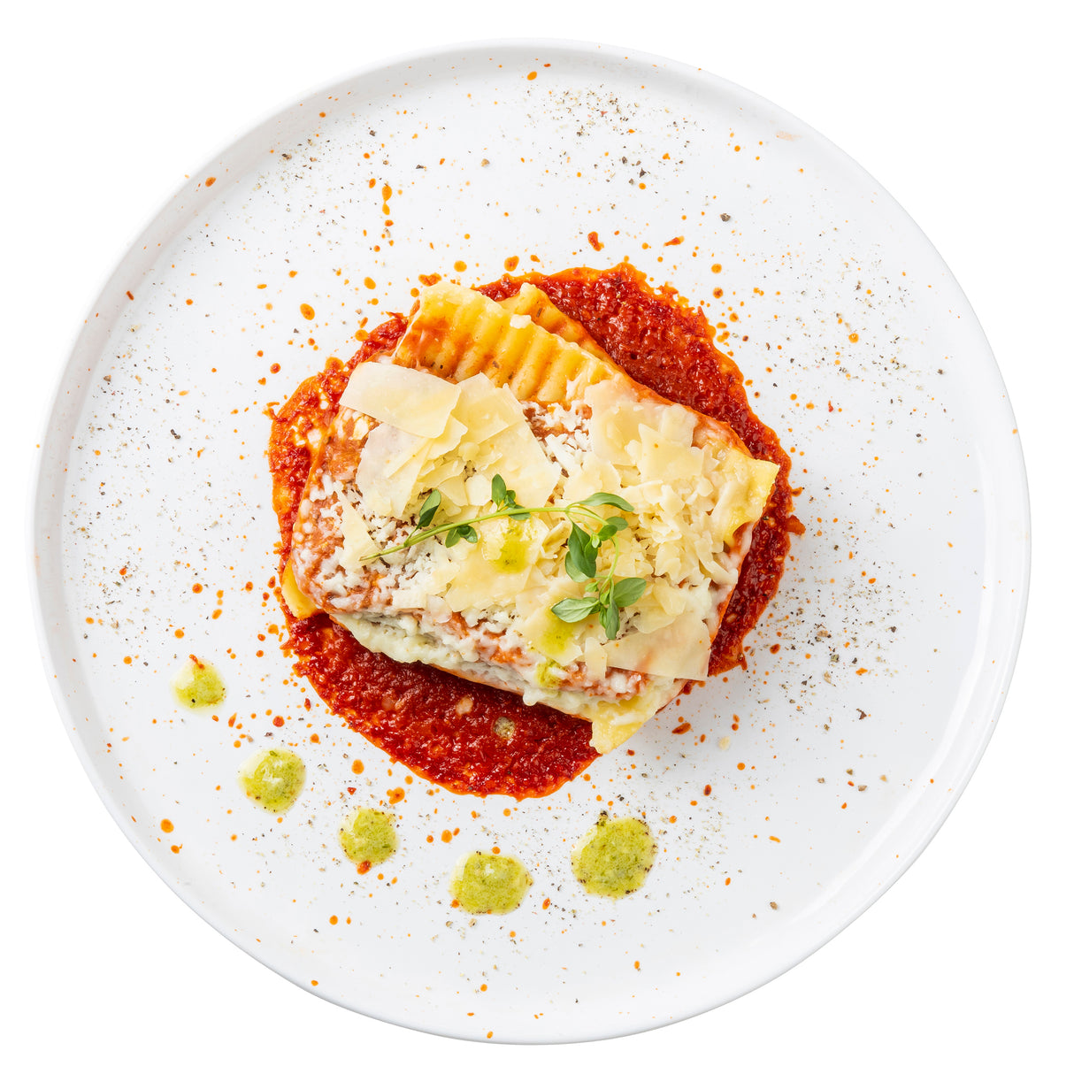 A square slice of lasagna topped with microgreens and shaved cheese sits on a red tomato sauce circle, garnished with green pesto dots and seasoning, all beautifully arranged on a white TableCraft melamine serving plate with scattered spice flecks.