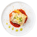 A square slice of lasagna topped with microgreens and shaved cheese sits on a red tomato sauce circle, garnished with green pesto dots and seasoning, all beautifully arranged on a white TableCraft melamine serving plate with scattered spice flecks.