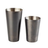 Two stainless steel cocktail shakers stand side by side. The taller, narrower shaker and the shorter, wider one both feature a brushed, gunmetal finish and are empty against a white background.