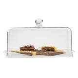 A rectangular clear glass TableCraft cake dome covers several pieces of layered dessert with a brown and yellow appearance, arranged on a matching rectangular cake plate. The dome has a round handle on top, set against a plain white background.