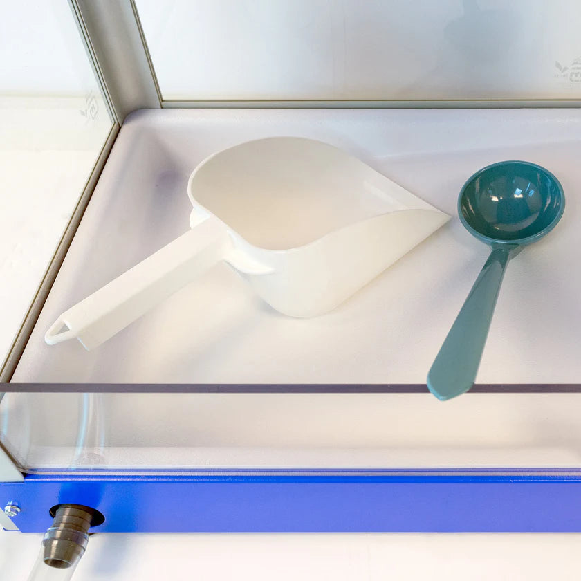 A white plastic scoop and a teal Sno pan ladle rest side by side on a clear, flat surface inside a glass display case with a blue frame, showcasing quality tools from Gold Medal Products.