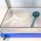 A white plastic scoop and a teal Sno pan ladle rest side by side on a clear, flat surface inside a glass display case with a blue frame, showcasing quality tools from Gold Medal Products.