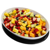 An oval white melamine serving platter from TableCraft Products, filled with assorted fresh fruit—sliced strawberries, cantaloupe, honeydew melon, pineapple chunks, and blueberries—sits on a black tray against a white background.