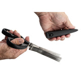 A person holds a pair of PerfectGrip herb scissors with black handles and five parallel stainless steel blades in one hand, and a black comb-like cleaning tool in the other, both against a white background.
