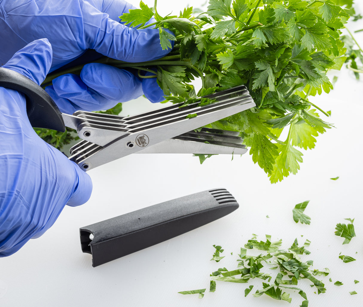 A person wearing blue gloves uses PerfectGrip herb scissors with stainless steel blades to chop fresh parsley, small green leaves falling onto a white surface. The black sheath for the scissors lies nearby.