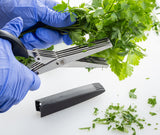 A person wearing blue gloves uses PerfectGrip herb scissors with stainless steel blades to chop fresh parsley, small green leaves falling onto a white surface. The black sheath for the scissors lies nearby.