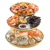 A three-tiered Seafood Tower holds oysters, shrimp, crab legs, mussels, scallops, and lobster on ice with lemon wedges. Cocktail sauce, ceviche, and a lemon half accompany the gold finish trays set against a white background.