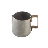 A small, metallic frothing cup with a matte gray finish and a wide brass handle. The pitcher, made from stainless steel, features a rounded body, a slight spout for pouring, and a simple, industrial design. The background is plain white.