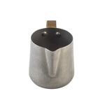 A stainless steel milk frothing cup with a brushed finish, curved spout, and sturdy rectangular handle. Photographed from above against a white background, the image highlights its circular opening and two rivets attaching the handle.
