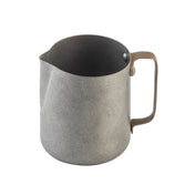 A gray stainless steel frothing cup with a matte finish, round body, and a small spout on one side. It features a sturdy brown handle attached with two rivets. The pitcher is shown empty against a white background.