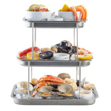 A 3-tier stainless steel Sharing Tower on ice displays cocktail shrimp with lemon, oysters, mussels, lobster tail, clams, crab legs, and dipping sauces. The seafood is neatly arranged with lemon wedges and small bowls of sauce on each tier.