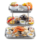 A three-tier Sharing Tower on stainless steel trays showcases shrimp, crab legs, lobster, oysters, clams, mussels, lemon wedges, dipping sauces, and ceviche with tortilla chips—all fresh and colorful for the perfect seafood experience.