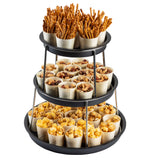 A 3-tier Sharing Tower with a black finish holds small wooden cups; the top tier has pretzel sticks, the middle has mixed nuts, and the bottom is filled with yellow popcorn. Each stainless steel tier neatly displays evenly arranged snack-filled cups.