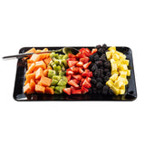 A rectangular black TableCraft platter displays neat rows of cubed cantaloupe, sliced kiwi, halved strawberries, blackberries, and cubed pineapple. A serving spoon rests at the top left corner. The white background highlights the colorful fruit arrangement.