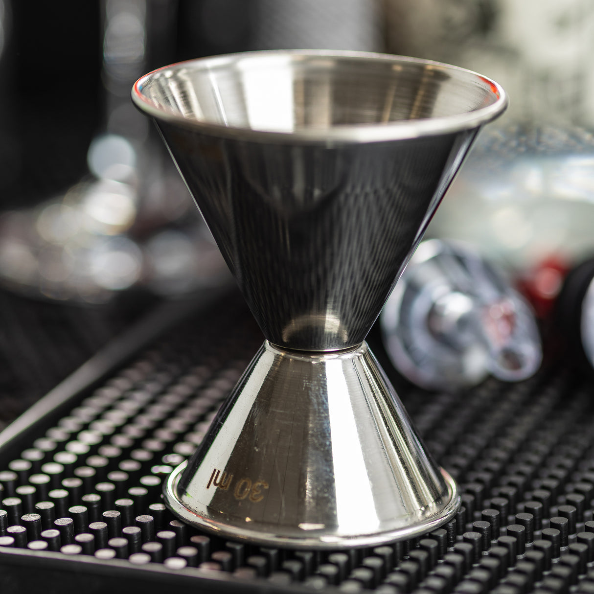 A close-up of a stainless steel jigger sits on a black bar mat. The 1 oz 2 oz jigger is marked with 30 ml, surrounded by blurred bar tools and bottles in the background, creating a professional bar setting.