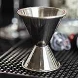 A close-up of a stainless steel jigger sits on a black bar mat. The 1 oz 2 oz jigger is marked with 30 ml, surrounded by blurred bar tools and bottles in the background, creating a professional bar setting.