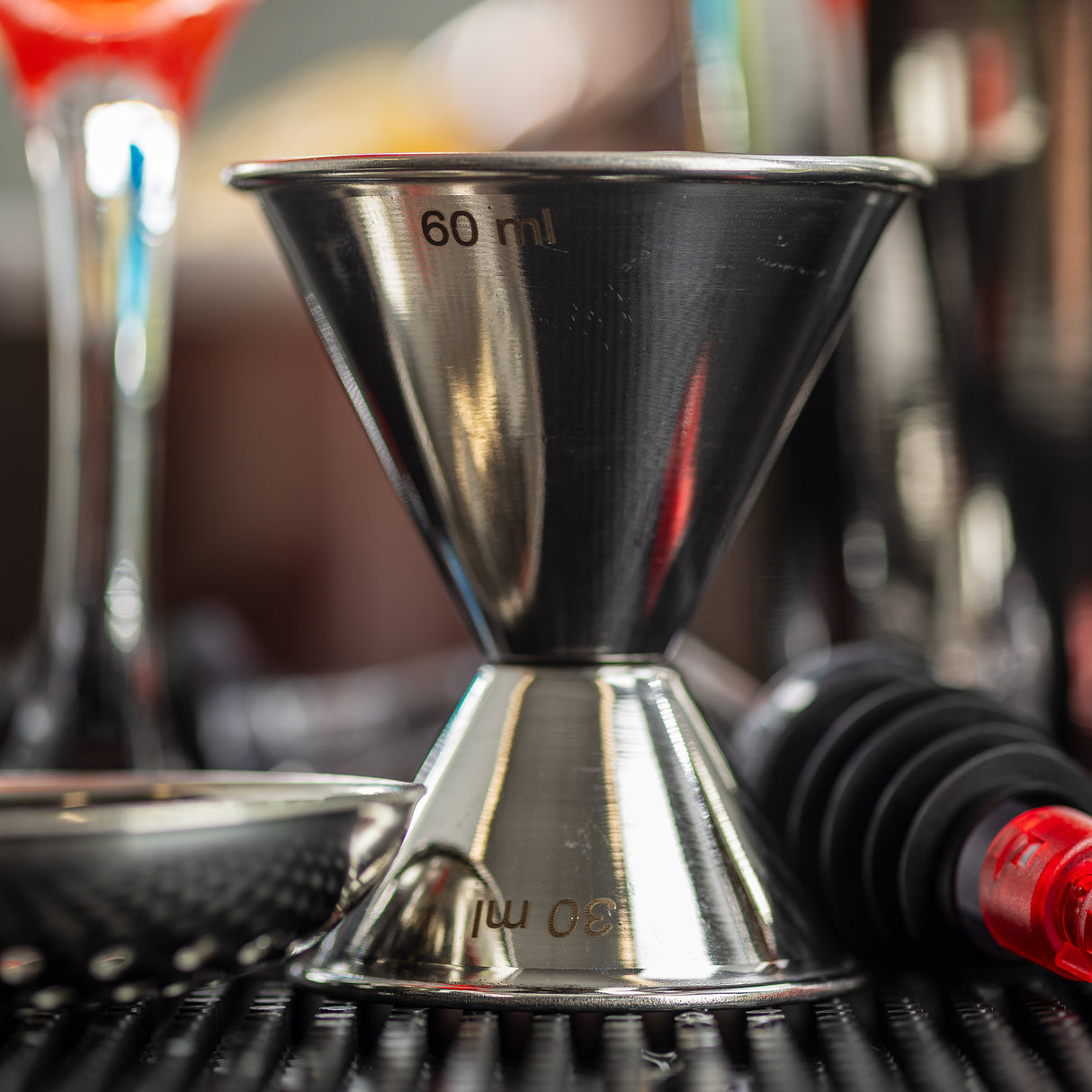 A close-up of a stainless steel jigger measuring cup, showing 60 ml and 30 ml markings. The 1 oz 2 oz jigger sits on a black bar mat with cocktail tools and blurred drinks in the shiny, reflective background.